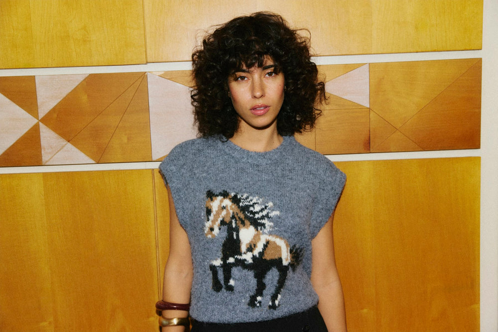 A model with curly dark hair wearing a gray knit sweater with a detailed horse graphic, standing in front of a yellow wood-paneled wall.